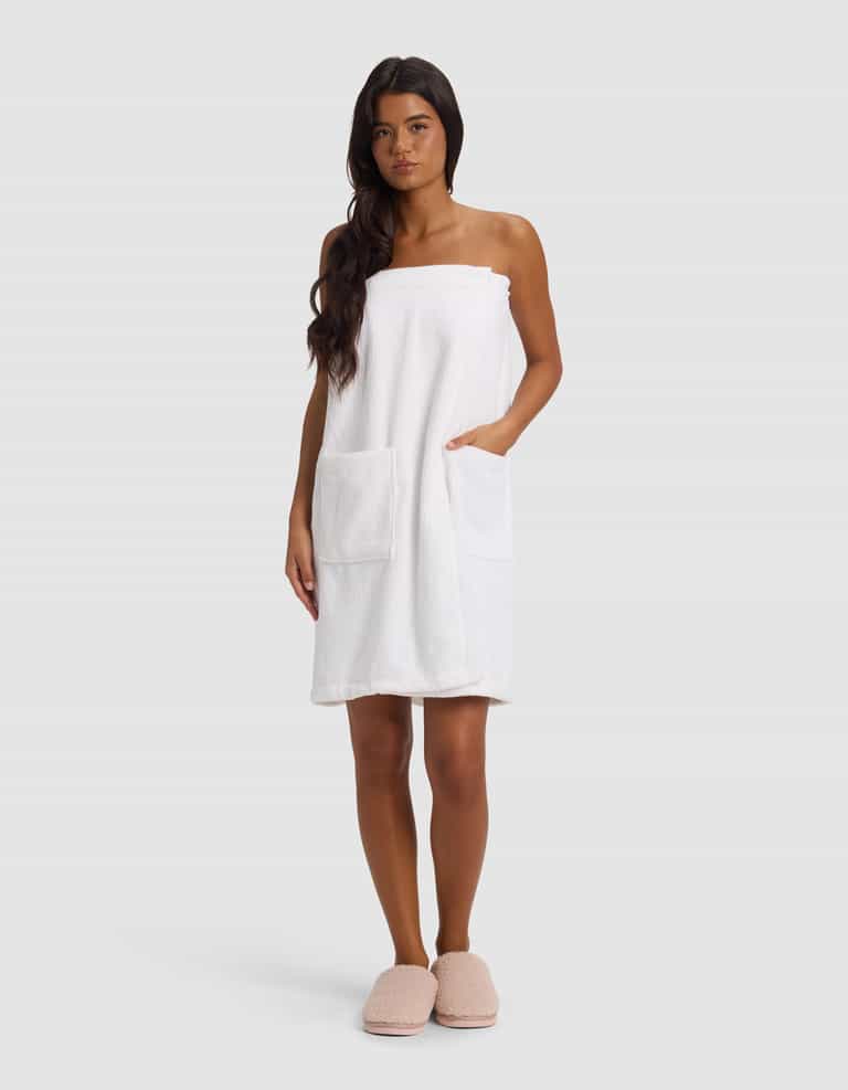 A woman with long dark hair stands facing forward, wearing the Cozy Earth Ribbed Terry Bath Wrap in white, featuring two large front pockets. She has a neutral expression and is set against a plain light gray background.|Color:White