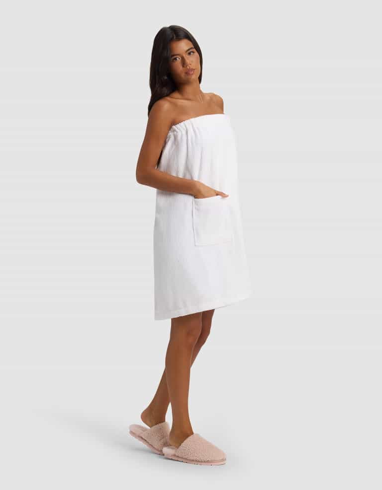 A woman with long dark hair models the Cozy Earth Ribbed Terry Bath Wrap, featuring a front pocket, paired with light pink slippers. She stands slightly turned against a plain light gray background.|Color:White