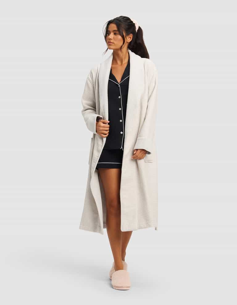 A woman wears the Cozy Earth Waffle Bath Robe over dark pajamas with white trim and matching slippers, her hair tied with a pink scrunchie as she gazes slightly to the side against a plain background.|Color:Light Grey