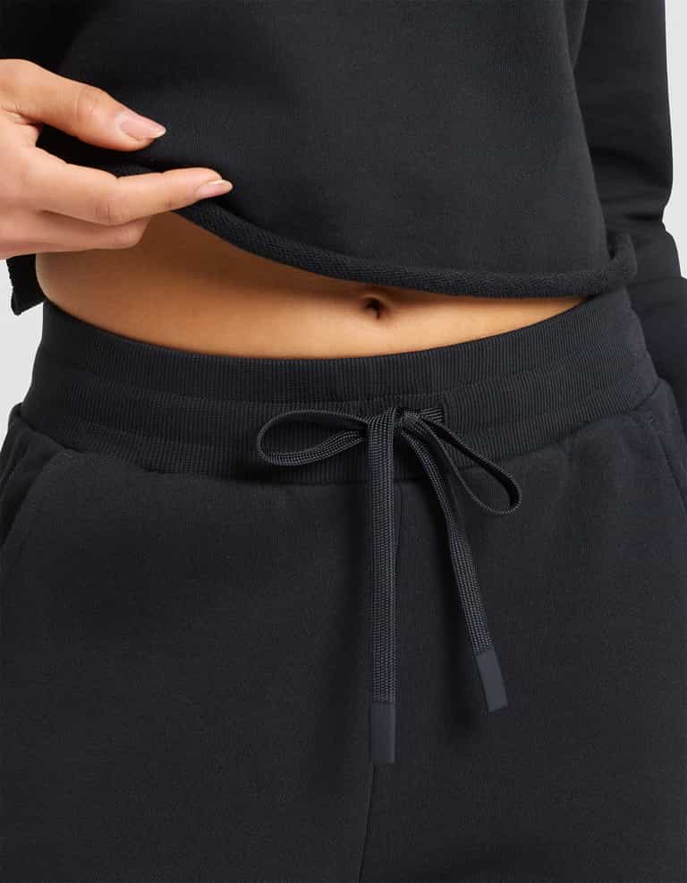 A woman models the Cozy Earth Women's Chelsea Cropped Pant in black, paired with a matching top that's lifted to reveal her bare midriff and navel. |Color:Jet Black