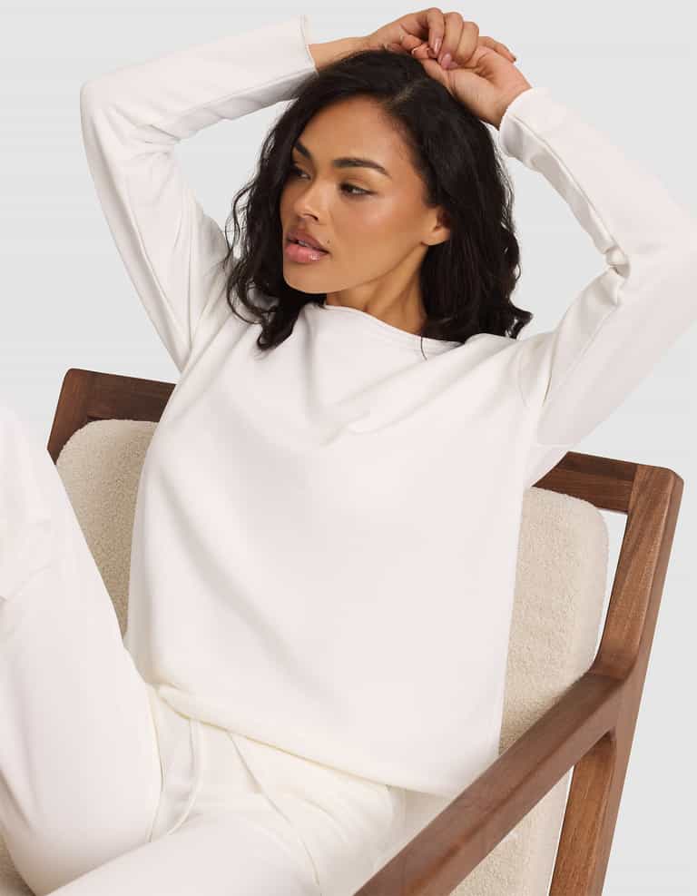 A woman with medium-length dark hair sits on a wooden chair, dressed in the Cozy Earth Women's Chelsea Pullover. With one arm raised and the other behind her head, she looks relaxed and gazes slightly to the side. |Color:Bone