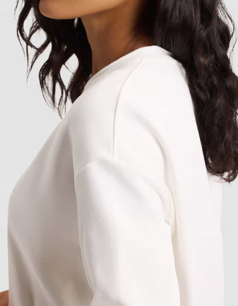 A woman with wavy dark hair wears the Cozy Earth Women's Chelsea Pullover, an off-white long-sleeve top, shown from the shoulders up and slightly turned to the side against a plain light background. |Color:Bone