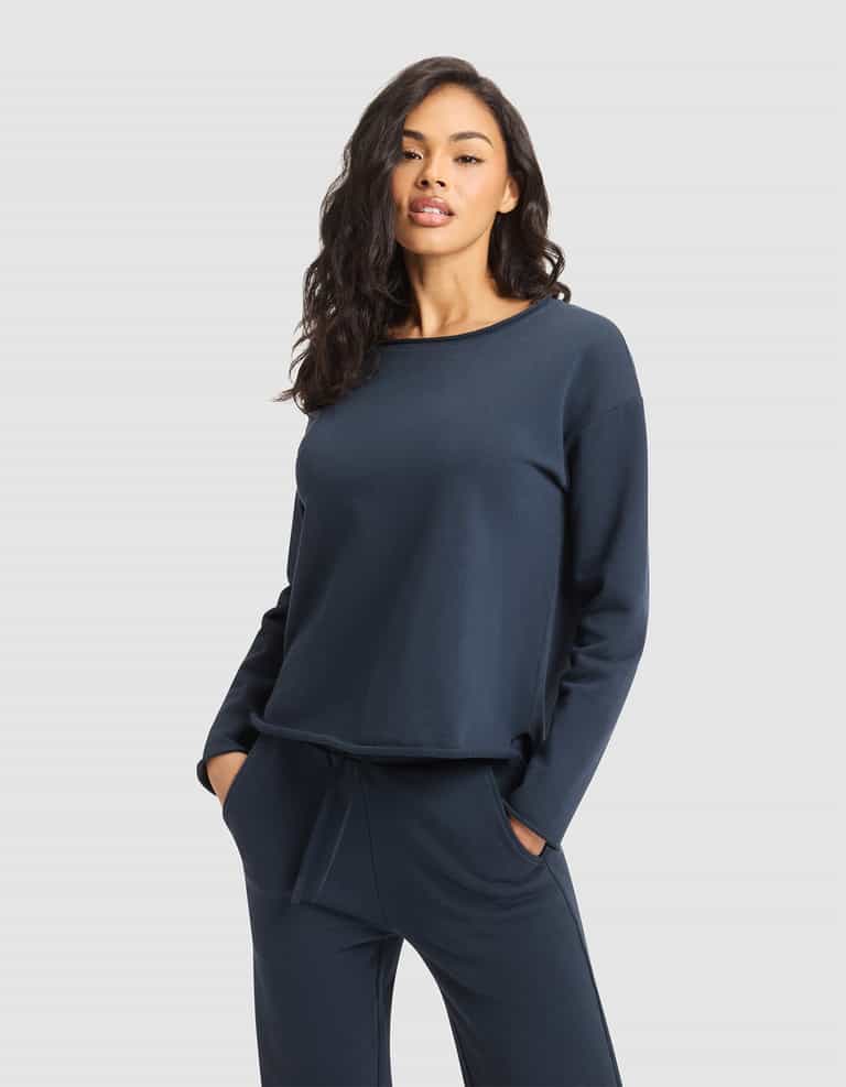 A woman with long, wavy dark hair wears the Cozy Earth Women's Chelsea Pullover and matching navy pants, standing with hands in her pockets against a plain light gray background. |Color:Eclipse
