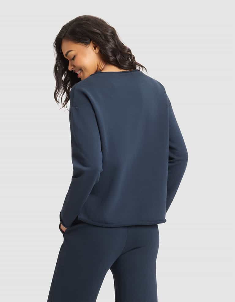 A woman with wavy dark hair, wearing the Cozy Earth Women's Chelsea Pullover in navy blue and matching pants, stands against a plain white background, facing away and smiling over her shoulder. |Color:Eclipse
