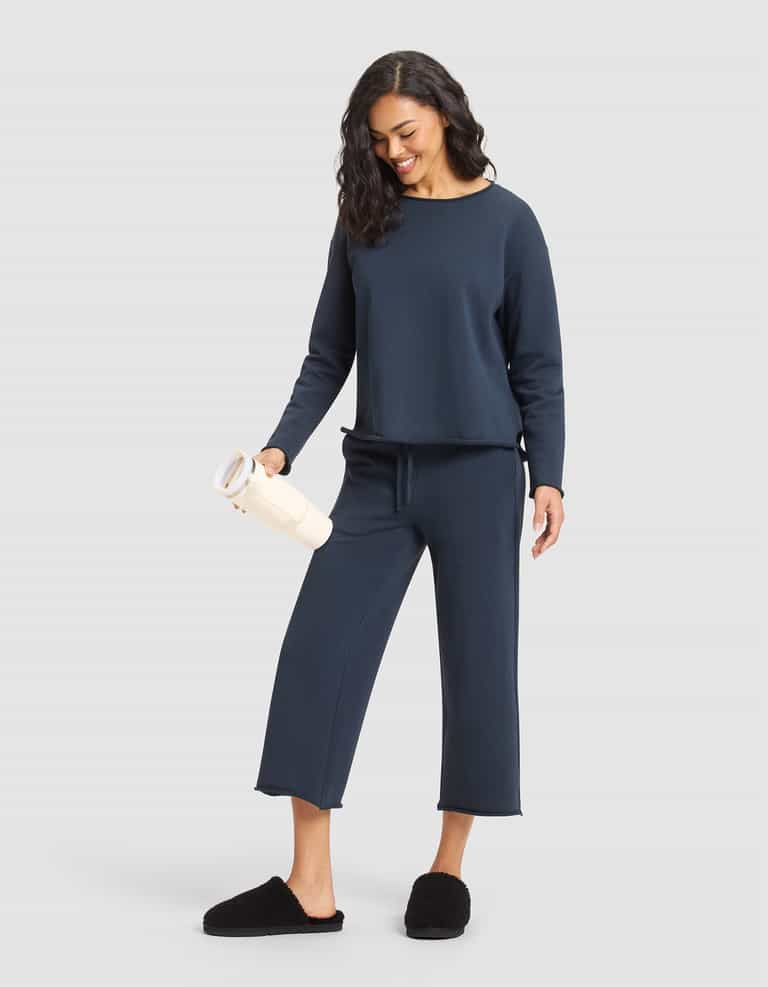 A woman wears the Cozy Earth Women's Chelsea Pullover in navy with matching wide-leg pants, paired with black slippers, and smiles while holding a light tumbler against a plain light gray background. |Color:Eclipse