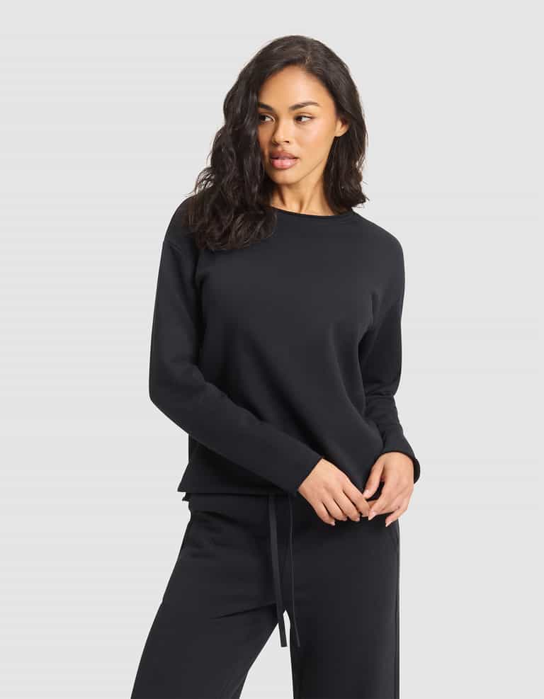 A woman with wavy dark hair models the Cozy Earth Women's Chelsea Pullover, a black long-sleeve sweatshirt, standing against a plain light gray background and looking slightly to her left. |Color:Jet Black
