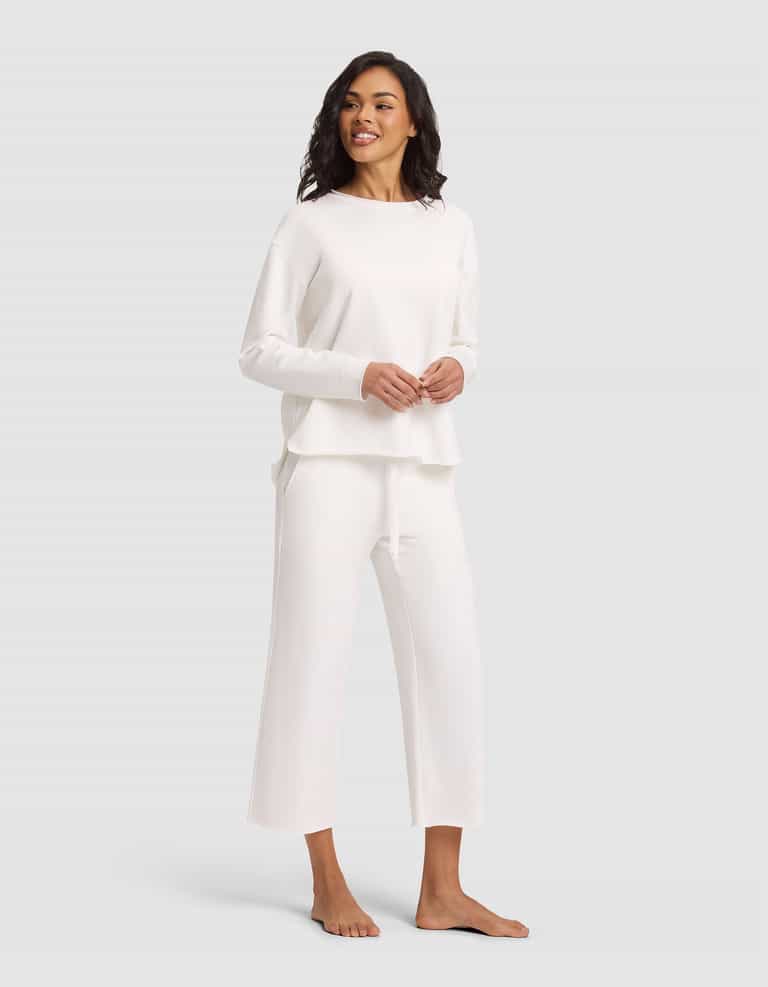 A woman stands barefoot, smiling, wearing the Cozy Earth Women's Chelsea Pullover—a loose-fitting, long-sleeve white top—paired with matching wide-leg pants against a plain light gray background. |Color:Bone