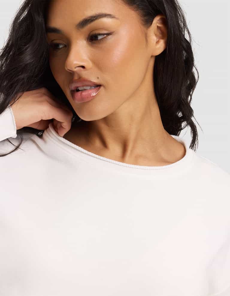 A woman with wavy dark hair and glowing skin, wearing the Cozy Earth Women's Chelsea Set, looks to the side while gently touching her neck. She is dressed in white against a neutral background. |Color:Bone