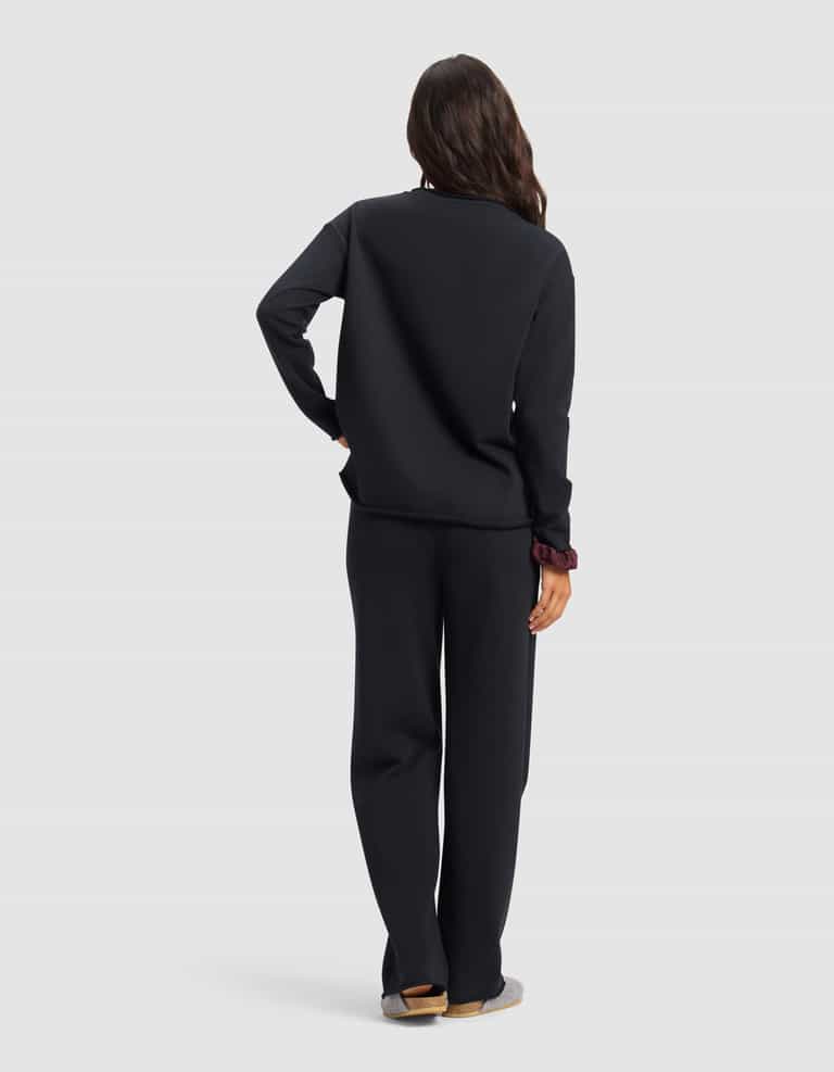 A person with long, wavy hair faces away, wearing the Cozy Earth Women's Chelsea Set—loose-fitting black sweatshirt and pants. One hand rests on their hip, slippers on their feet. The background is plain light gray.|Color:Jet Black