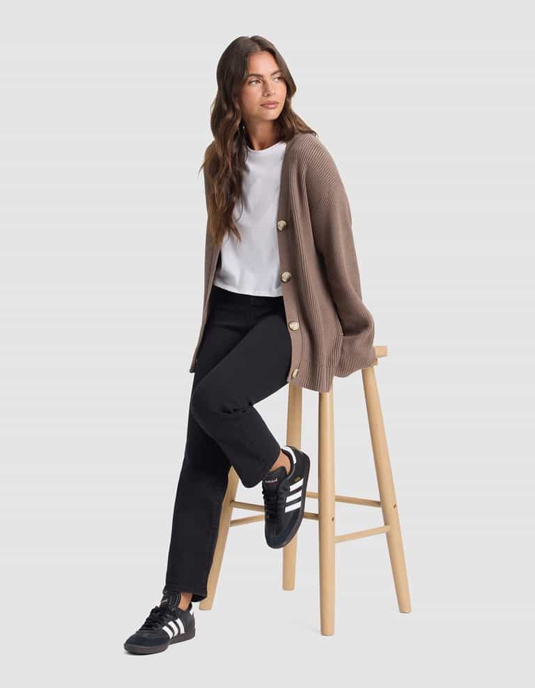 A woman with long brown hair, wearing Cozy Earth's Oversized Classic Cardigan in taupe, a white t-shirt, black jeans, and black Adidas sneakers, sits on a wooden stool against a plain light gray background.|Color:Ebony