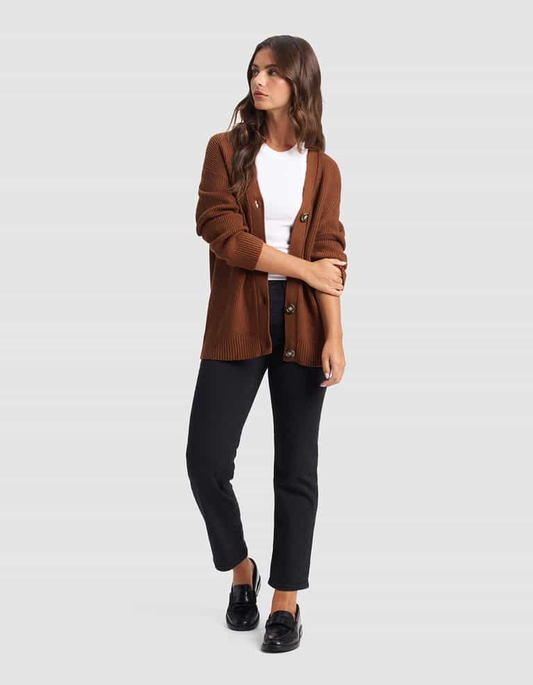 A woman stands against a plain background wearing the Cozy Earth Oversized Classic Cardigan over a white tee, black pants, and black loafers. She has long hair, looks slightly to the side, and folds one arm.|Color:Spice