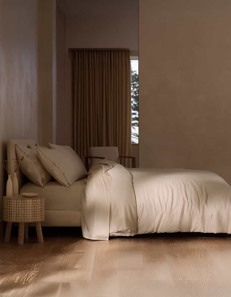 A minimal, modern bedroom features a neatly made bed with Cozy Earth's Bamboo Duvet Cover in cream, a round nightstand with a vase of dried plants, wood floors, and a window with brown curtains partially revealing greenery outside.