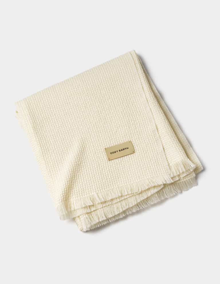 A folded, cream-colored Alpaca Waffle Throw with a textured design, fringe edges, and a rectangular "Cozy Earth" label, displayed on a plain white background.|Color:Creme