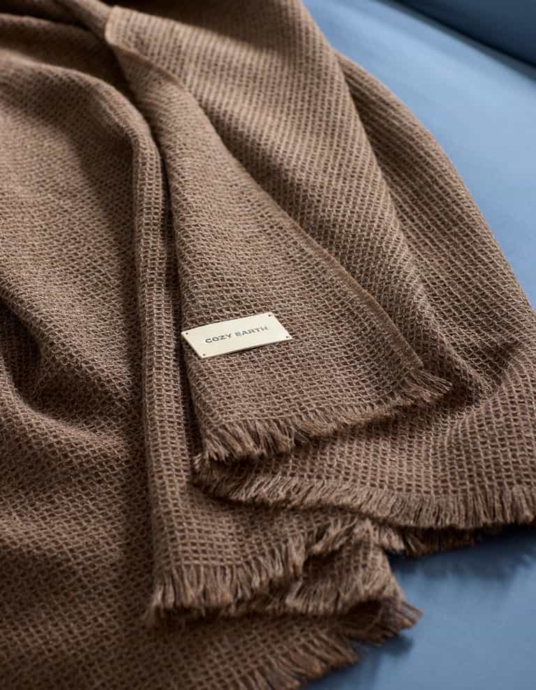 A brown Alpaca Waffle Throw by Cozy Earth, featuring a textured weave and fringed edges, is draped over a blue surface. A small white label with the brand name "Cozy Earth" is visible on the blanket.|Color:Ebony