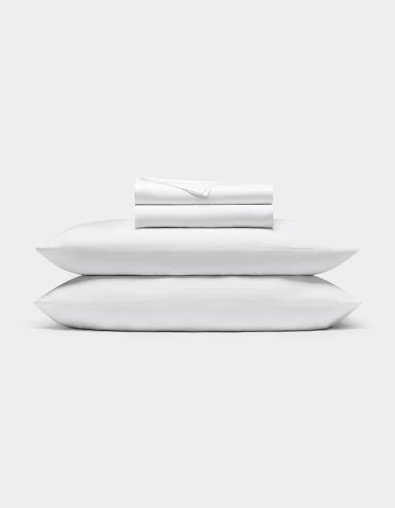 A neatly folded Bamboo Sheet Set by Cozy Earth is stacked on two white pillows, all set against a plain light gray background.|Color:White
