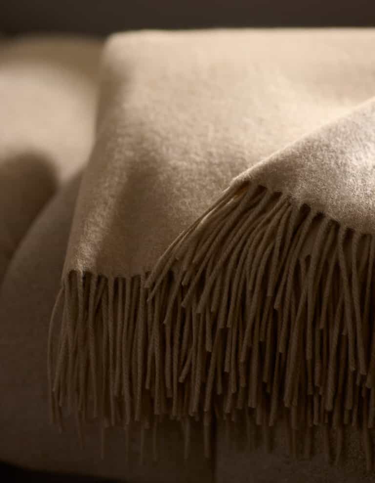 A close-up of the Cozy Earth Cashmere Tassel Throw, a soft beige blanket with tassel edges, neatly folded atop another in a warmly lit setting.|Color:Dune