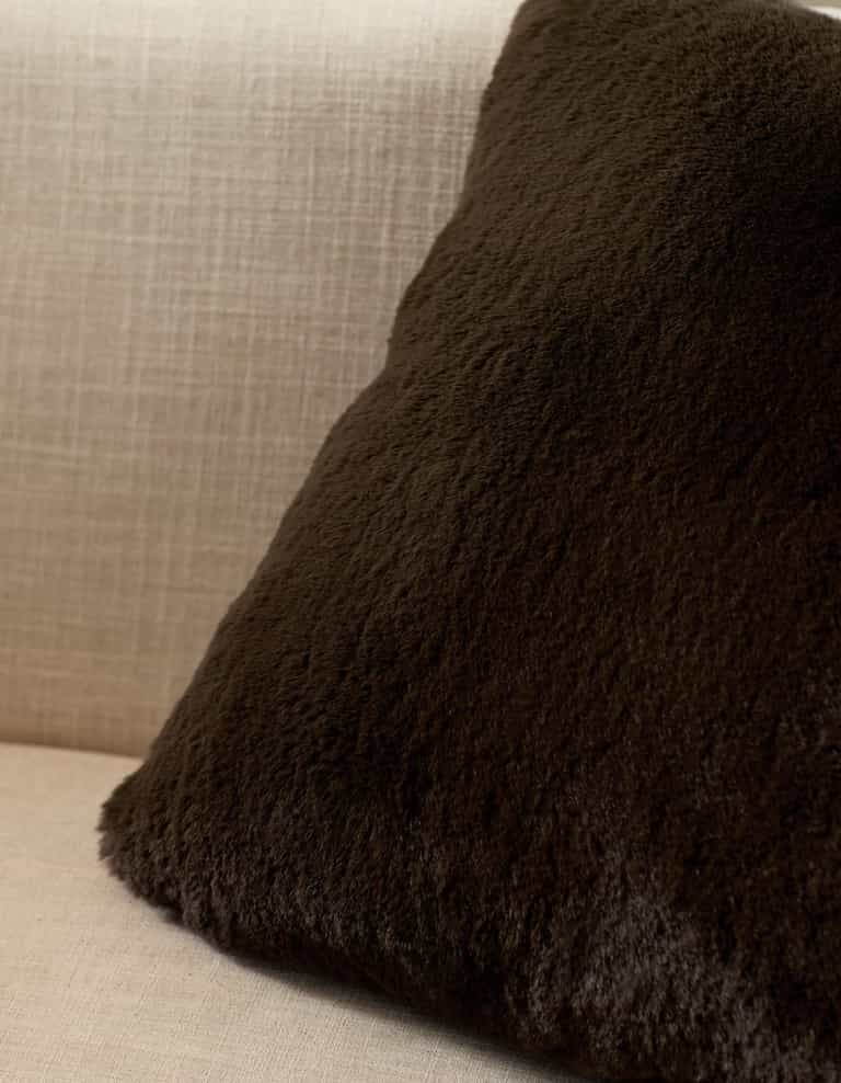 A Cozy Earth Cuddle Pillow in dark brown rests against the back of a light beige, textured fabric sofa.|Color:Espresso
