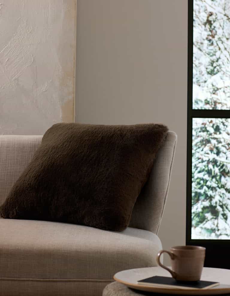 A beige sofa with the Cozy Earth Cuddle Pillow in dark brown sits by a window overlooking snow, while a ceramic cup and saucer rest on a small table nearby.|Color:Espresso