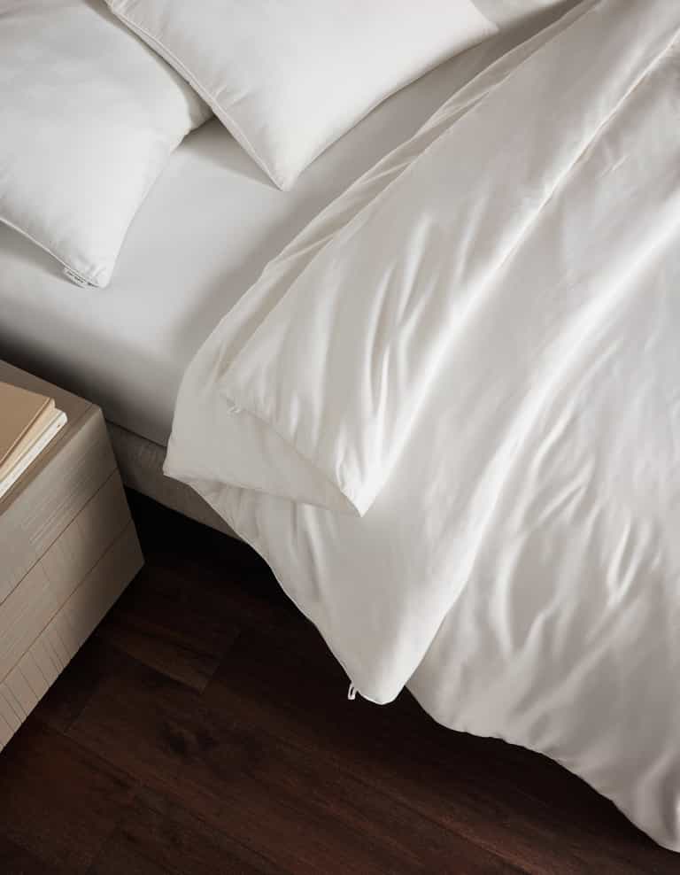 A bed dressed in Cozy Earth’s Silk Comforter, paired with white pillows, stands beside a light wood nightstand with two closed books—all atop a dark wood floor.Coal Stripe/Striped Alabaster, Laurel Stripe/Striped Bone