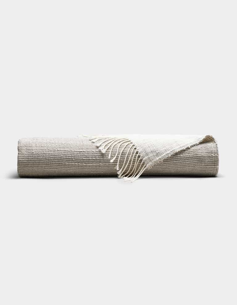 The Cozy Earth Baja Tassel Throw, a striped blanket in neutral beige and off-white tones with fringed edges, is neatly rolled and displayed on a plain, light background.|Color:Grey