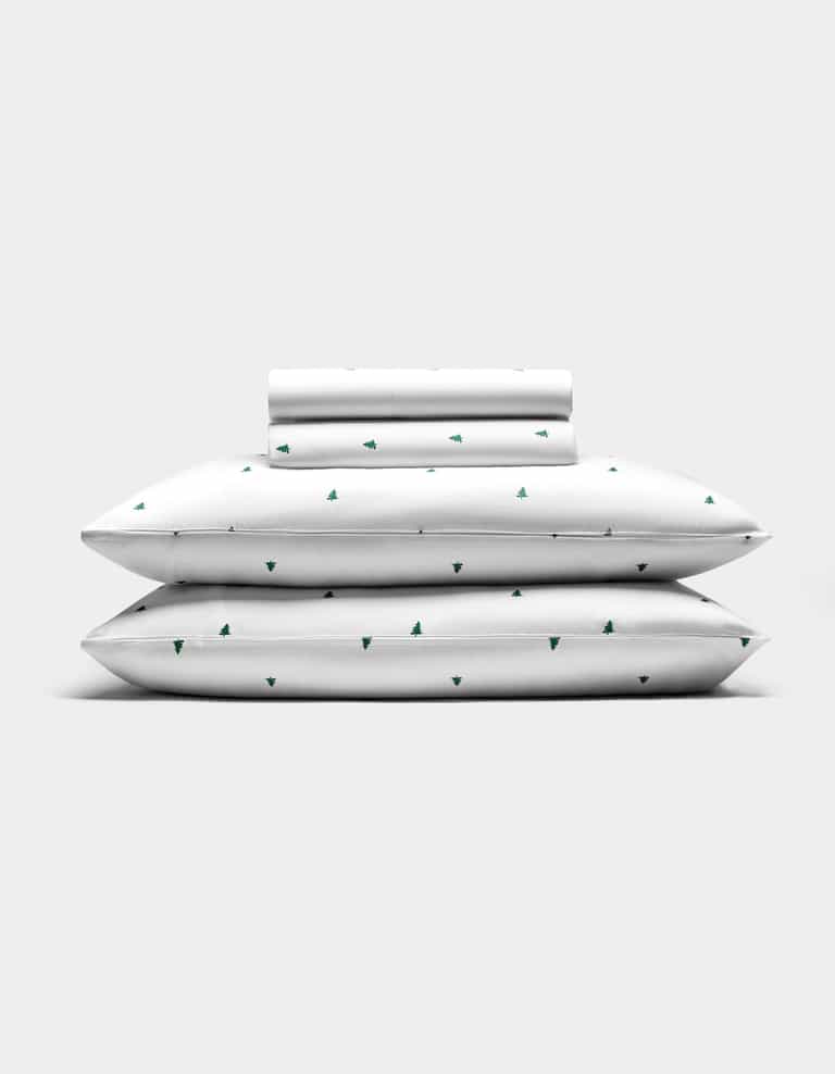 The Cozy Earth Bamboo Sheet Set features two pillows, a folded sheet, and a rolled pillowcase—all white with small green tree patterns—arranged against a plain light gray background.|Color:Jade Tree Snow