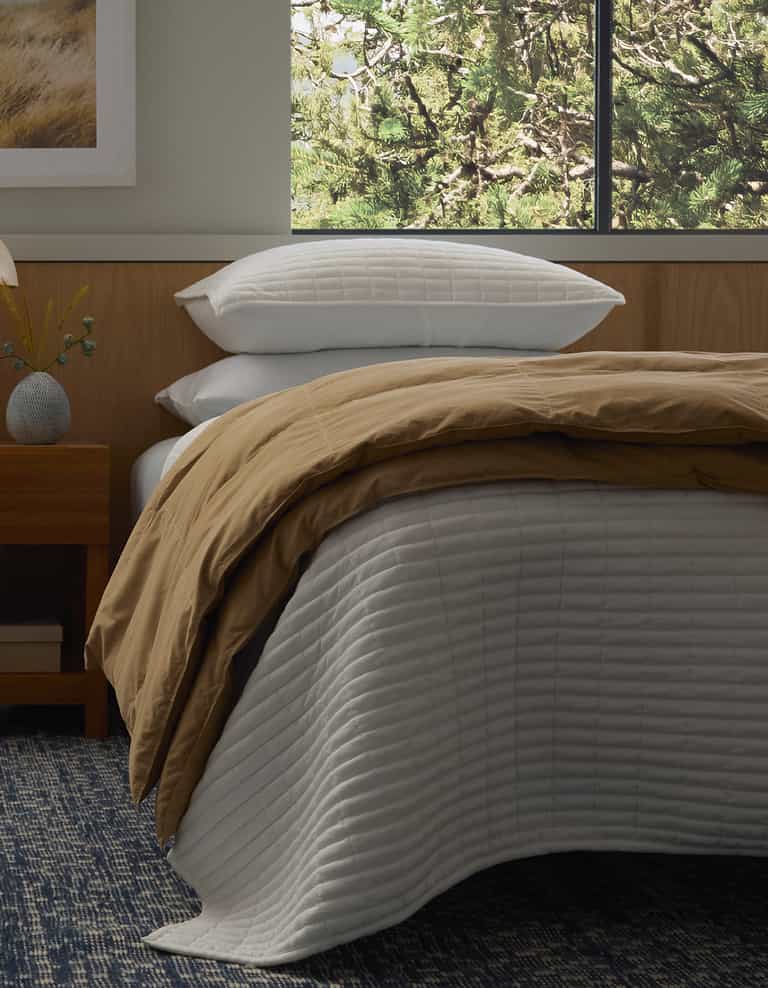A bed styled with Cozy Earth's Down Alternative Throw features white quilted bedding, a tan comforter, and four stacked white pillows. Next to it sits a wooden nightstand with a vase and lamp, while green tree branches are visible through the window.|Color:Sandalwood