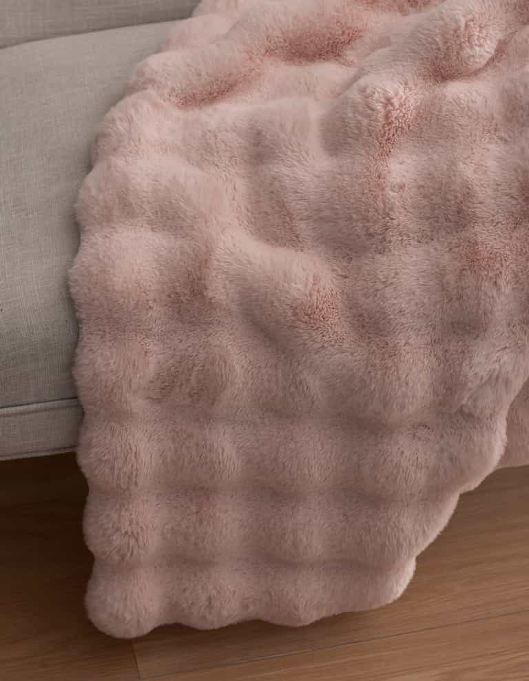 The Cozy Earth Bubble Cuddle Blanket in soft light pink faux fur is draped over the edge of a light gray sofa, cascading gently onto a wooden floor.|Color:Blossom