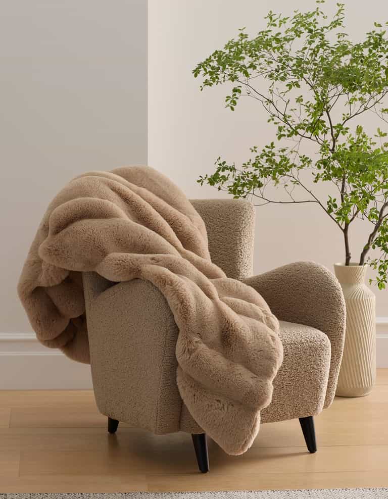 A soft, beige armchair with black legs is draped with the plush Channel Cuddle Blanket by Cozy Earth. Nearby, a leafy green plant in a textured white vase sits on a wooden floor against a light-colored wall.|Color:Brandy