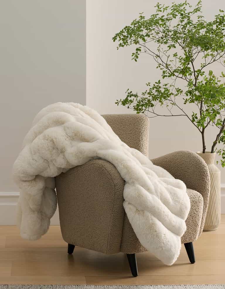 A taupe armchair with the Cozy Earth Channel Cuddle Blanket draped over it sits on a light wood floor beside a green potted plant and a light-colored wall.|Color:Creme