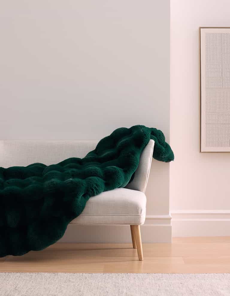 A modern, light gray bench with wooden legs is draped with the Cozy Earth Double Bubble Cuddle Blanket in dark green. The bench sits against a pale wall, beside a framed minimalist artwork on a hardwood floor.|Color:Adrift