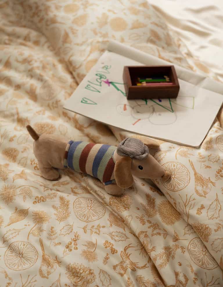 A plush dachshund toy in a striped sweater and hat rests on a bed made with the Cozy Earth Bamboo Duvet Cover; colored pencils and a child’s drawing on white paper are nearby.|Color:Winter Toile Dijon
