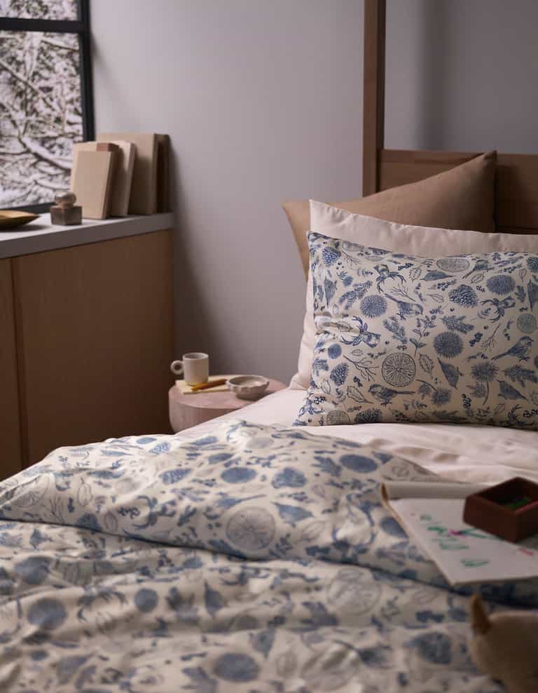 A cozy bedroom features a bed topped with the Cozy Earth Bamboo Sheet Set, beige pillows, a small bedside table holding a cup and saucer, books, and a window displaying a snowy scene outside.|Color:Winter Toile Pacific