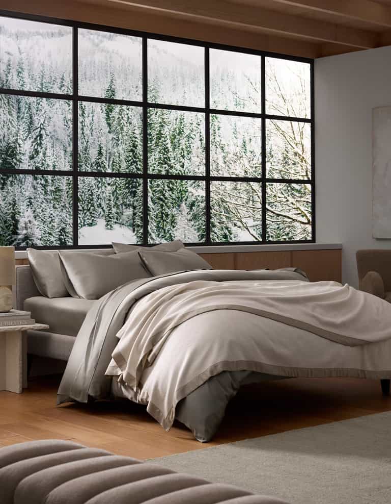 A modern bedroom features Cozy Earth's Bamboo Blanket in neutral tones on an unmade bed, large windows with snowy forest and mountain views, wooden floors, minimalist furniture, and abundant natural light for a serene, cozy feel.|Color:Oat/Driftwood