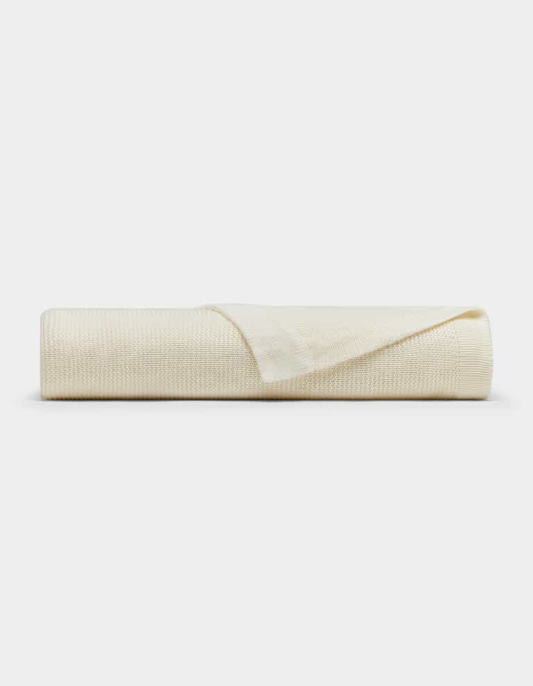 The Cozy Earth Cloud Knit Blanket by Cozy Earth is cream-colored with a textured pattern, neatly rolled and displayed on a white background. One edge is folded back, highlighting the blanket's thickness and texture.|Color:Ivory