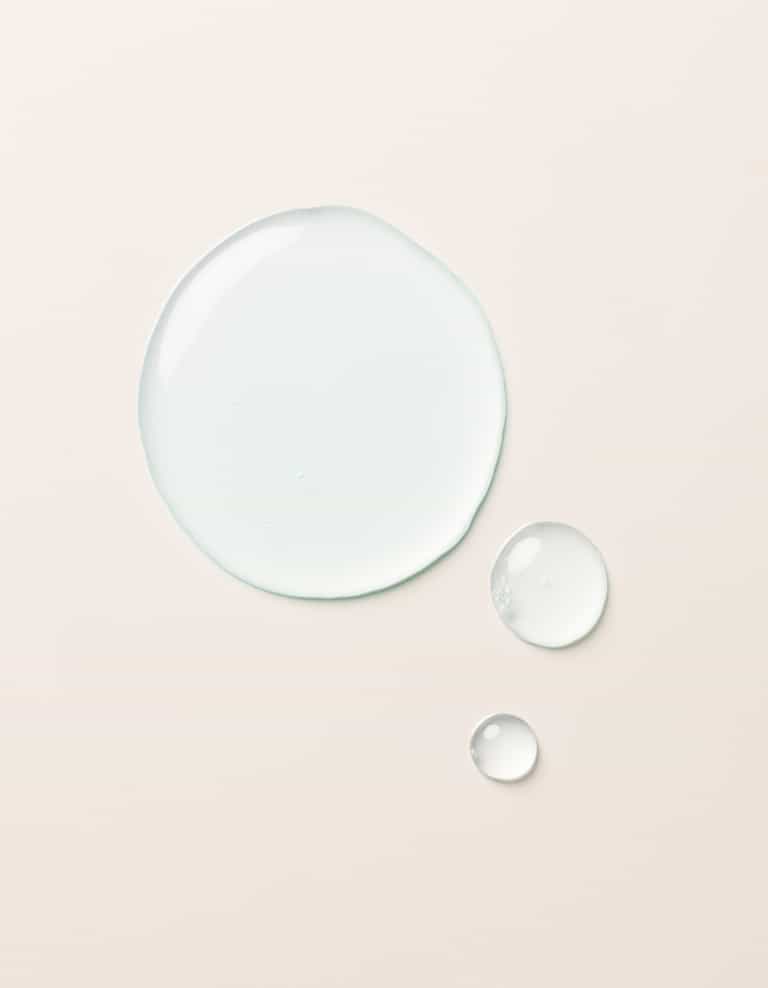 The Hydration Duo by Cozy Earth is shown as a large drop and two smaller drops of clear, light blue liquid on a smooth, light beige surface. The drops vary in size but all maintain a rounded shape, appearing glossy and slightly reflective.