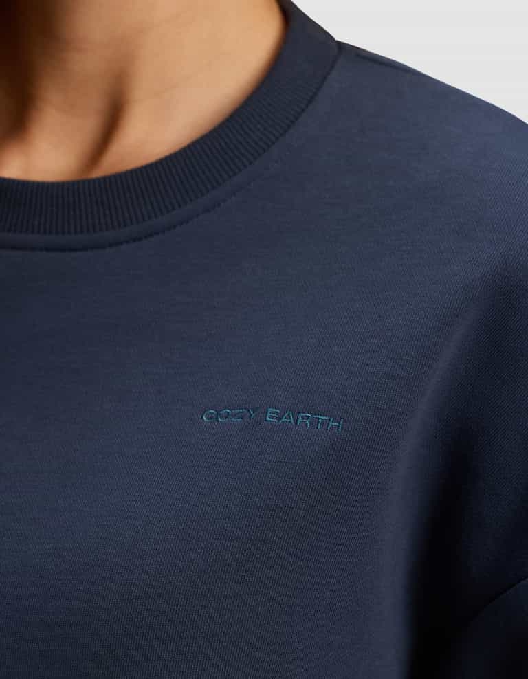Close-up of a person wearing the Women's Cityscape 2.0 Crewneck by Cozy Earth in navy blue, featuring small light blue "Cozy Earth" embroidery on the chest. The collar and soft fabric texture are clearly visible.|Color:Eclipse