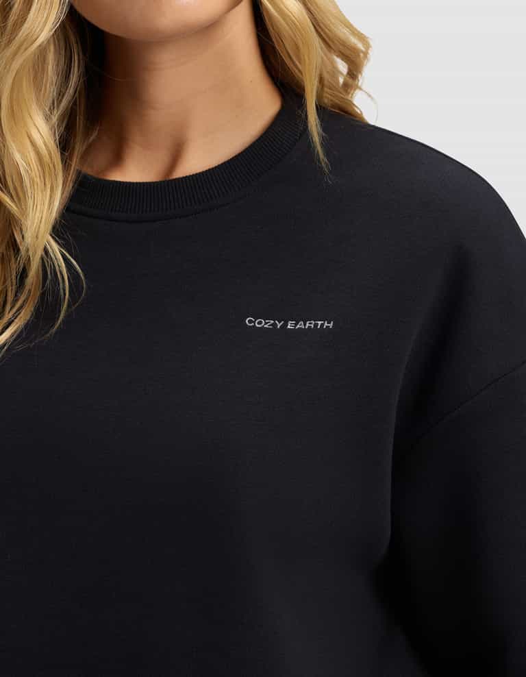 A close-up shows a person in the Women's Cityscape 2.0 Crewneck by Cozy Earth, featuring "COZY EARTH" embroidered in small white letters on the black chest; their blonde hair is visible, with most of their face out of frame.|Color:Jet Black