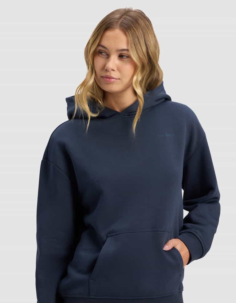 A woman with long, wavy blonde hair wears the Cozy Earth Women's Cityscape 2.0 Hoodie in dark navy, with one hand in the pocket, gazing slightly to the side against a plain light gray background.|Color:Eclipse