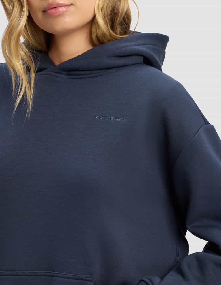 A woman wears the Cozy Earth Women's Cityscape 2.0 Hoodie in navy blue, featuring a small embroidered logo on the chest. Only her lower face and upper body appear against a plain background.|Color:Eclipse