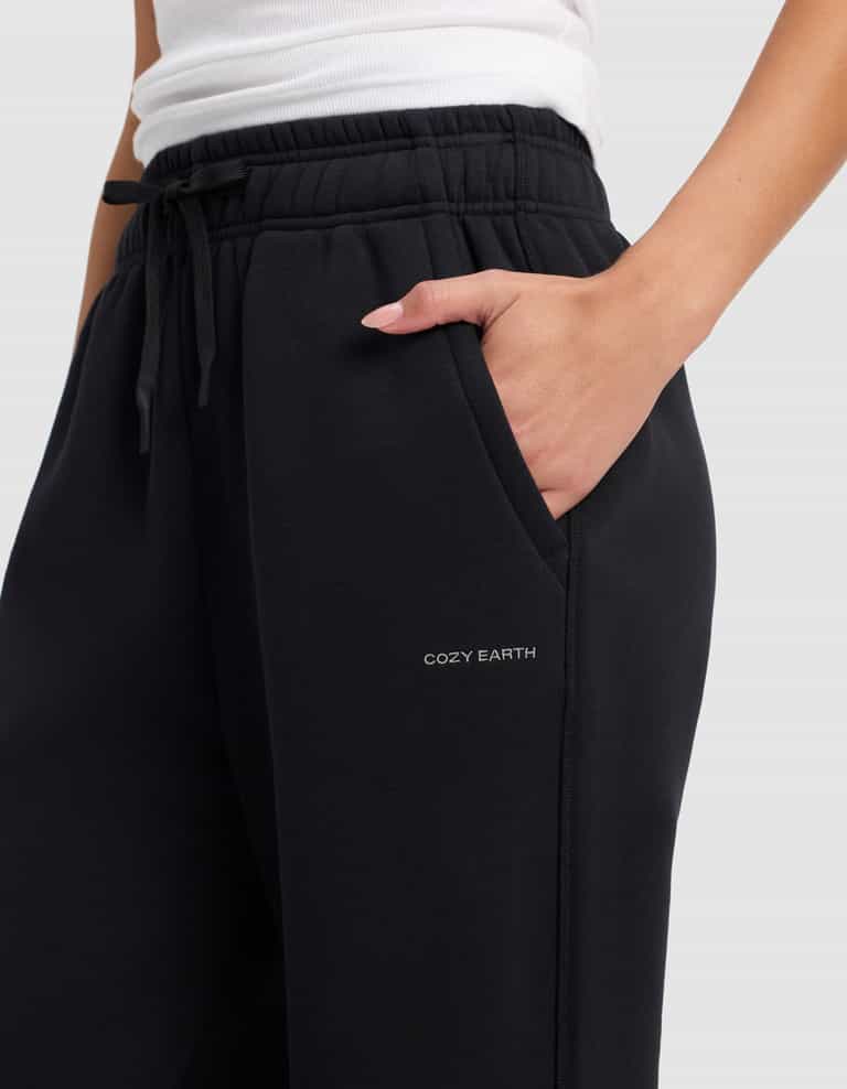 A person wears Cozy Earth Women's Cityscape 2.0 Wide Leg Pant in black, featuring "COZY EARTH" printed near the pocket, with one hand in the pocket and a white top.|Color:Jet Black