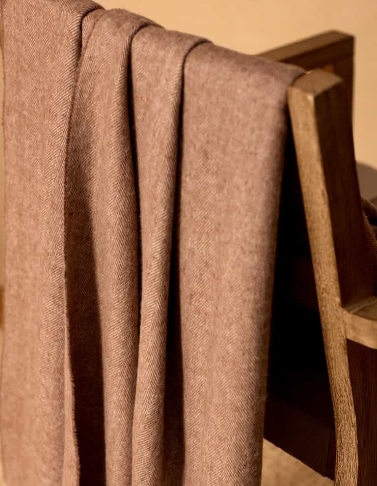 The Cozy Earth Bamboo Herringbone Tassel Throw, in light brown with a herringbone pattern, is draped neatly over a wooden chair, bathed in warm lighting.|Color:Truffle