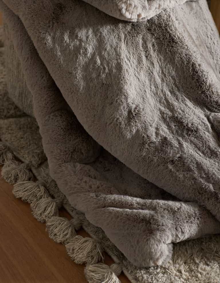 A close-up of the Cozy Earth Cuddle Blanket, a soft gray faux fur throw, draped over a textured beige rug with fringed edges on a wooden floor.|Color:Dove Grey