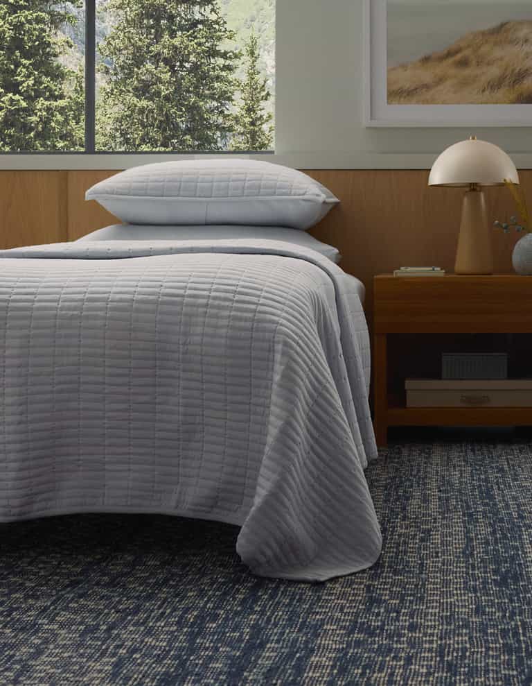 A bed with the Cozy Earth Bamboo Jersey Quilt in light gray and matching pillow is next to a wooden nightstand with a lamp, all atop a blue patterned rug. A large window and a framed landscape art piece are visible in the background.|Color:Shore