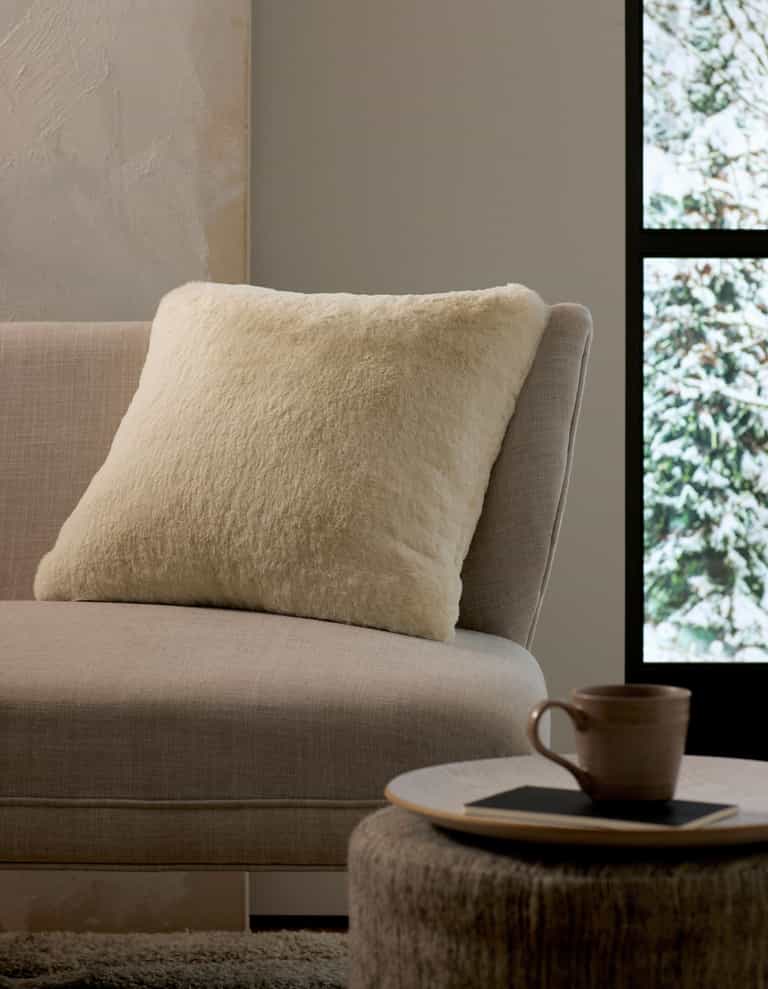 A cozy interior scene with a light beige sofa, a fluffy Cozy Earth Cuddle Pillow in cream, a round ottoman holding a cup and saucer, and a window framing a snowy outdoor view.|Color:Creme |Size:20" x 20"