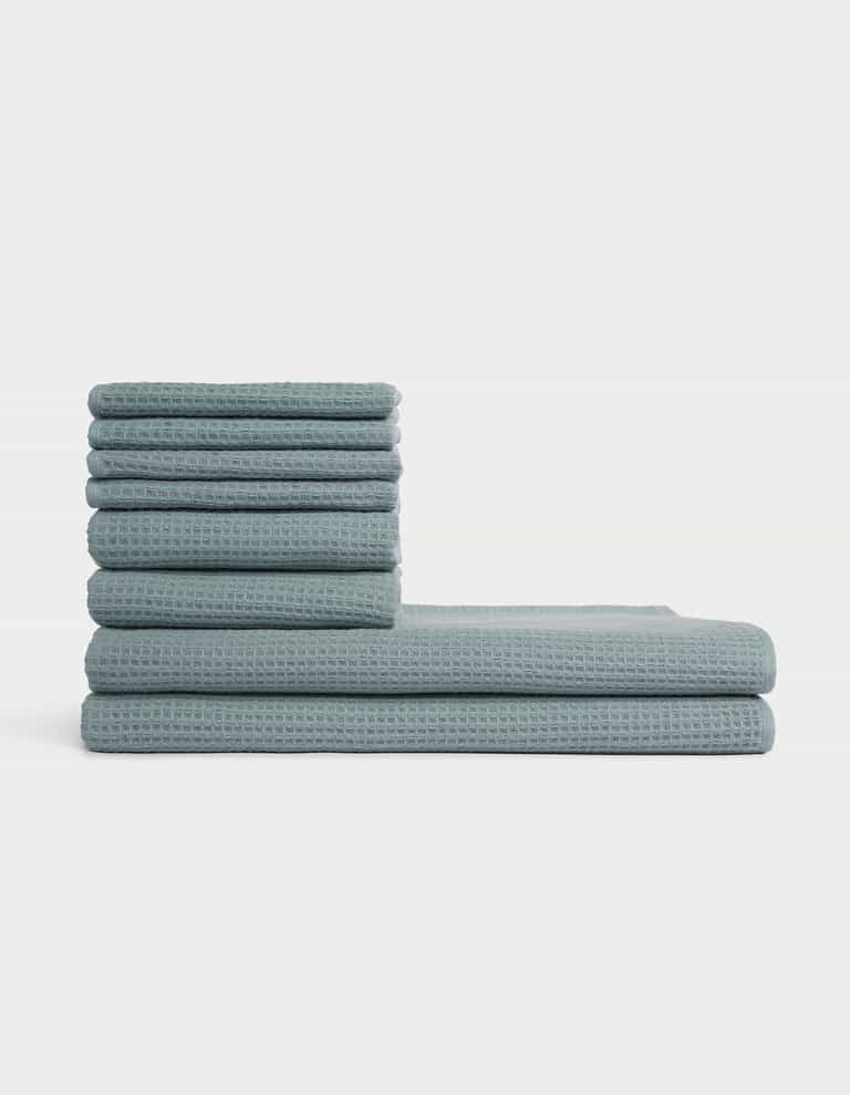 The Cozy Earth Waffle Bath Towel Set features blue, waffle-textured towels stacked in two vertical piles of varying heights against a plain white background. |Color:Brook