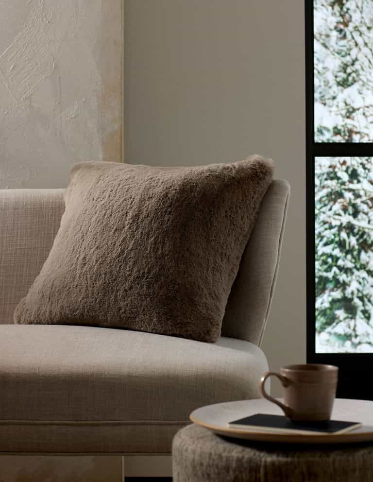 A cozy, neutral-toned living space features the Cozy Earth Cuddle Pillow in taupe on a beige sofa next to a round side table with a ceramic mug and book, framed by a window view of snowy trees.|Color:Walnut