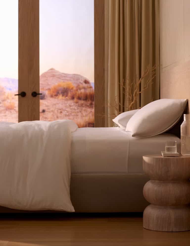 A bed made with Cozy Earth's Bamboo Sheet Set features crisp white linens beside a wooden nightstand with a glass of water and bottle. Large windows reveal a scenic desert landscape, while neutral tones create a calm, minimalist vibe. |Color:White