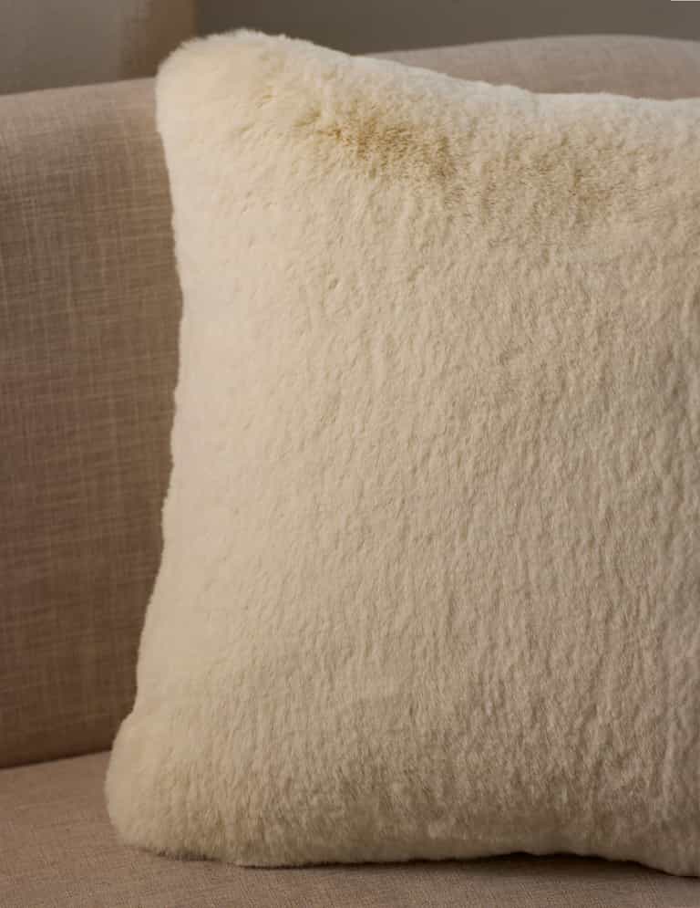 A close-up of the Cuddle Pillow by Cozy Earth, featuring a fluffy cream color and soft texture, placed on a light beige fabric sofa.|Color:Creme |Size:20" x 20"