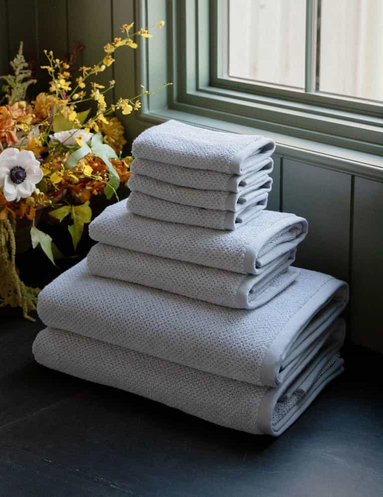 The Cozy Earth Nantucket Bath Towel Set, in varying sizes, is neatly stacked on a dark surface near a window, with yellow and white flowers in the background. |Color:Heathered Harbor Mist