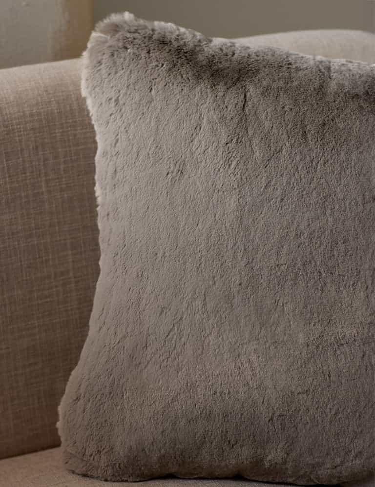 The Cozy Earth Cuddle Pillow, a large square in light gray faux fur, rests against the back of a beige fabric sofa.|Color:Dove Grey |Size:20" x 20"